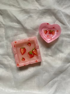 two pink plates with strawberries in them on a white cloth covered bed sheet, one is shaped like a heart and the other is shaped like a rectangle