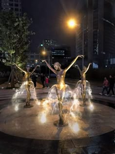 three statues in the middle of a fountain with lights shining on them and people standing around