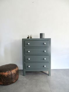 a small chest of drawers next to a stool in a room with white walls and concrete flooring