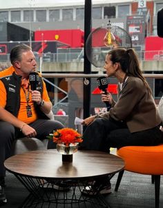 two people sitting on orange chairs talking to each other in front of a tv screen