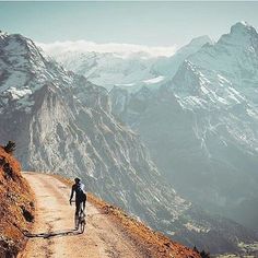 a person riding a bike down a dirt road in the mountains with snow capped peaks