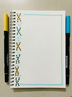 an open notebook with cross stitch on the cover and two pens sitting next to it