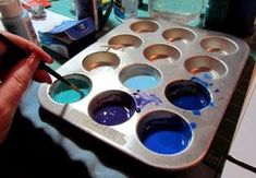 a person holding a paintbrush in front of a muffin tin filled with blue liquid