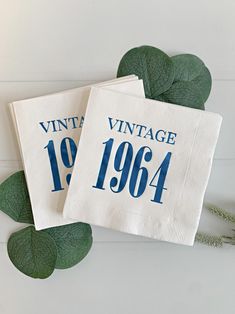 two white napkins with blue numbers on them and green leaves next to each other