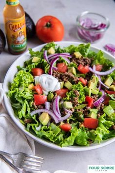 a salad in a bowl with dressing on the side next to an avocado and tomatoes