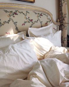 an unmade bed with white linens and floral wallpaper on the headboard