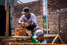 two men are working on building bricks in front of a brick wall, and one man is holding out his hand to another man