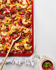 a red casserole dish with shrimp, peppers and sausage on it next to a wooden spoon