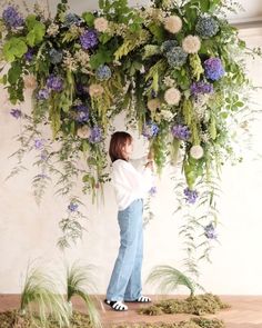 a woman standing in front of a flower covered wall with blue and white flowers hanging from it's sides