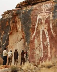 three men are standing in front of a rock wall with an image of a human being painted on it