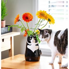 a black and white cat standing next to a vase with flowers in it on a table