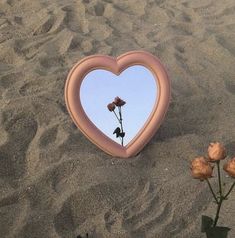 a pink heart shaped mirror sitting on top of a sandy beach next to two flowers