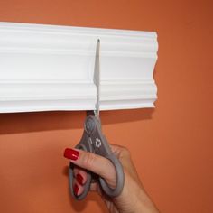 a hand holding a pair of scissors in front of an orange wall with white trim