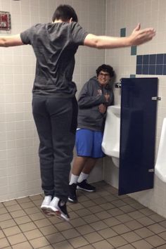 two men standing in a bathroom with urinals on the wall and one holding his arms out