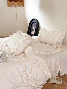 an unmade bed with white ruffles and pillows on it, next to a clock