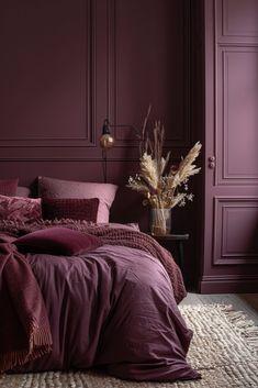 a bed with purple sheets and pillows in front of a dark colored wall, next to a vase with dried flowers