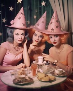 three women wearing pink hats sitting at a table with food and drinks in front of them