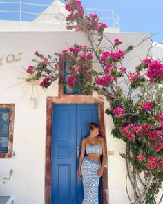 a woman standing in front of a blue door with pink flowers growing on the side