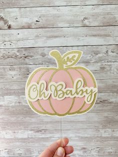 a hand holding up a pink and gold pumpkin cutout with the word oh baby on it