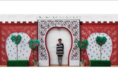 a man standing in front of a red and white wall decorated with trees, bushes and flowers