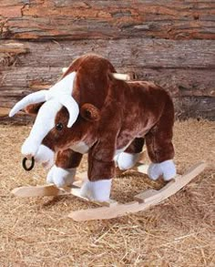 a brown and white stuffed cow sitting on top of a wooden rocking horse in hay