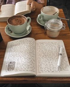 an open book sitting on top of a wooden table next to two cups of coffee