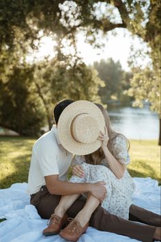 a man and woman sitting on the ground with hats over their heads, in front of trees