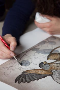 a woman is drawing on paper with a red pen and pencil while another person holds a white piece of paper in front of her