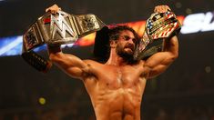 a shirtless man holding two wrestling belts in front of his head and shoulders with one arm raised