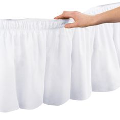 a hand is pulling up the side of a white bed skirt with pleated edges