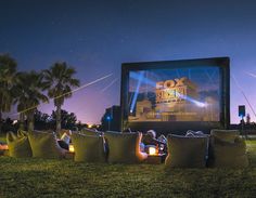 an outdoor movie theater with people sitting in the grass watching it on screen at night