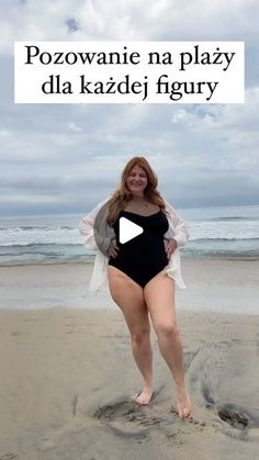 a woman in a black swimsuit standing on the beach with her arms behind her back