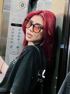 a woman with red hair and glasses standing next to a metal wall holding a cell phone