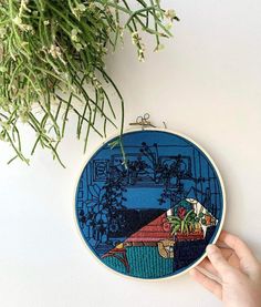 someone is holding up a cross - stitch pattern in front of a potted plant