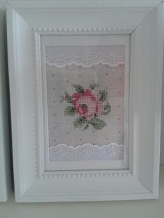 a white frame with a pink rose on it and lace around the edges, in front of a wall