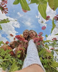 two women are taking a selfie with flowers in the foreground and clouds in the background