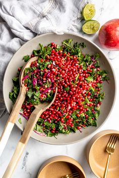 a salad with pomegranate and greens in a bowl next to gold utensils