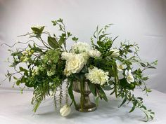 a vase filled with white flowers and greenery on a white tableclothed surface