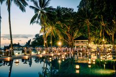 many candles are lit in the water at night near some palm trees and tables with lights on them