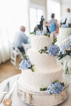 a white wedding cake with blue flowers and figurines on top is sitting on a table