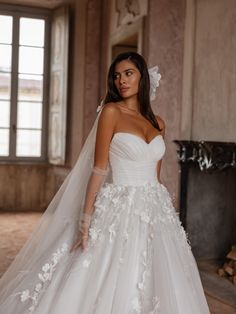 a woman wearing a wedding dress with flowers on the skirt and veil, standing in front of a fireplace