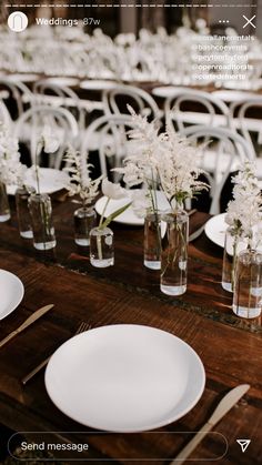 the table is set with white flowers in vases and plates on it, along with silverware