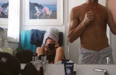 a shirtless man takes a selfie in the mirror while his friend looks at him