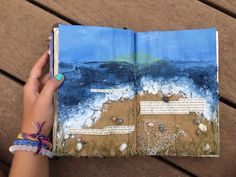 a hand holding an open book on top of a wooden table next to the ocean