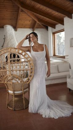 a woman in a white dress standing next to a wicker chair and looking at the camera