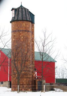 a red barn and silo in winter with snow on the ground, trees and fence