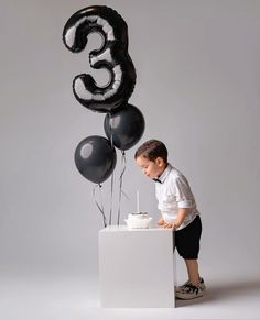 a young boy standing in front of a birthday cake with black balloons and the number three on it