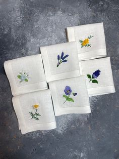 six embroidered napkins with flowers on them sitting on a gray table top, one is white and the other is blue