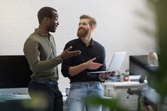 two men standing in an office talking to each other and holding laptops with their hands