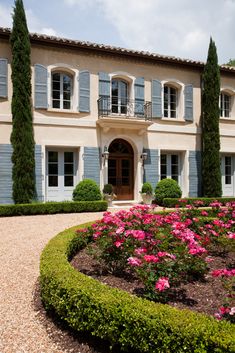 a large house with blue shutters and pink flowers in the front yard, on a sunny day